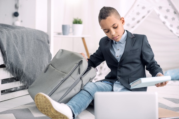 Schulsachen. Trauriger afroamerikanischer Junge, der auf Boden sitzt, während Buch von Schultasche erhält