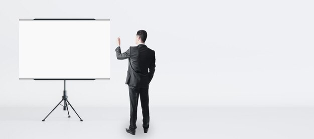 Schulungs- und Geschäftsseminarkonzept mit Mann im schwarzen Anzug, Rückansicht vor leerem weißem Flipchart mit Platz für Ihr Logo oder Text auf hellem Wandhintergrund