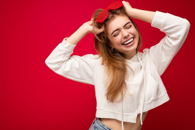 Schuss der schönen glücklichen lächelnden jungen blonden Frau lokalisiert über der roten Wand, die lässigen weißen Kapuzenpulli und stilvolle Sonnenbrille genießt und Spaß hat. Speicherplatz kopieren