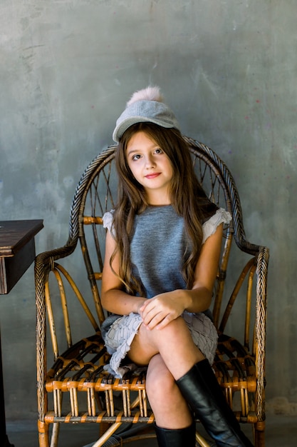 Schuss des schönen brünetten kaukasischen kleinen Mädchens, das graues Kleid und mit einem Hut trägt, die Kamera mit entzückendem Lächeln betrachtet, auf einem hölzernen Sessel sitzend. Glückliches Kindheitskonzept