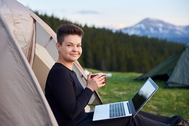 Schuss einer glücklichen jungen frau, die ihr getränk hält und sich im freien beim campen in der nähe des zeltes mit ihrem laptop entspannt
