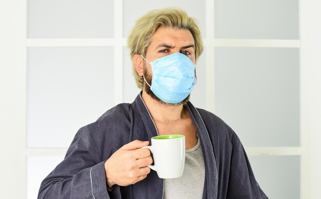 Schutz vor Coronavirus Mann trägt Gesichtsmaske Virus, das durch Tröpfchen und engen Kontakt übertragen wird Das Tragen einer Maske schützt vor Coronavirus Kerl in Maske trinkt Tee oder Kaffee zu Hause Quarantänekonzept