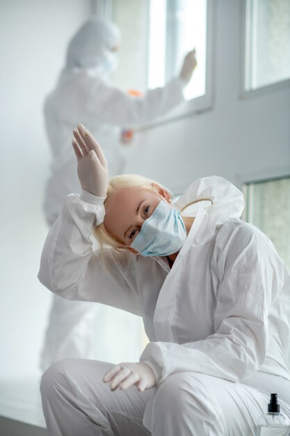 Schutzmaßnahmen. Müde blonde Ärztin in Schutzkleidung und medizinischer Maske sitzt neben dem Fenster und ihre Kollegin desinfiziert Scheiben