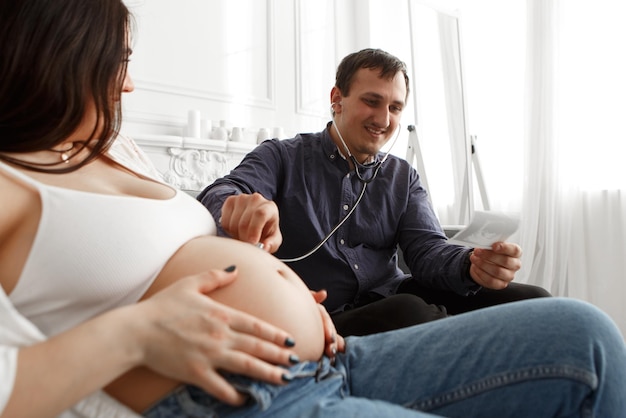 Schwangere Frau bei Arztbesuch hält Ultraschallfoto, hört Bauch mit Stethoskop