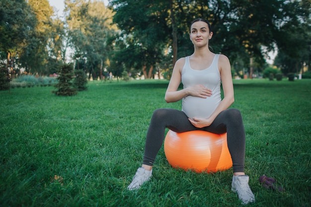 Schwangere Frau der Junge sitzen auf orange Eignungsball im Park. Sie hält die Hände um den Bauch. Frau schauen gerade.