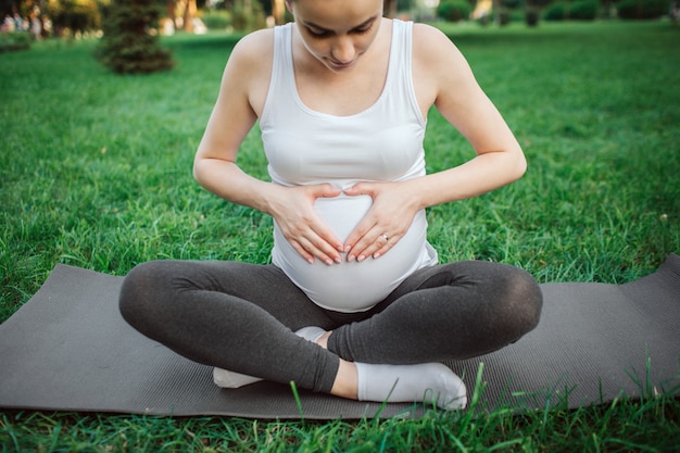 Schwangere Frau der Junge sitzen in der Lotoshaltung auf Yogakameraden im Park. Sie schaut auf den Bauch und hält dort die Hände in Herzform.