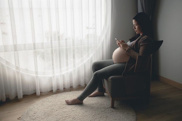 Schwangere Frau des jungen Asiaten, die Handy im Wohnzimmer verwendet.