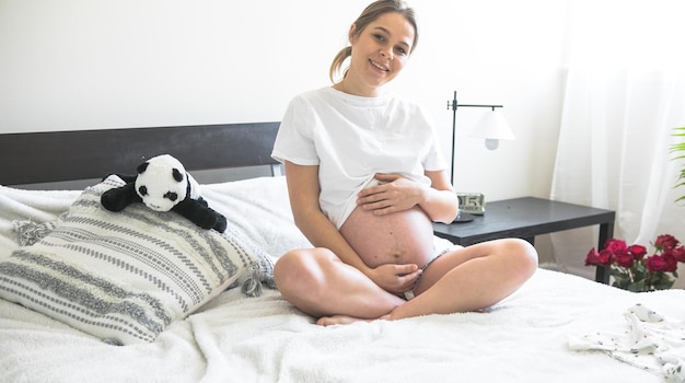 Schwangere Frau, die auf einem Bett sitzt und das Konzept der Schwangerschaft, Mutterschaft und Schwangerschaftsvorsorge hält Mutter mit einem neuen Leben