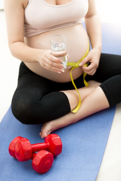 Schwangere Frau, die Bauch mit Maßband auf Fitnessmatte misst und ein Glas Wasser hält