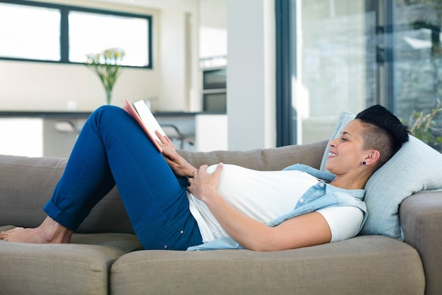 Schwangere Frau, die ein Buch beim Lügen auf Sofa im Wohnzimmer liest