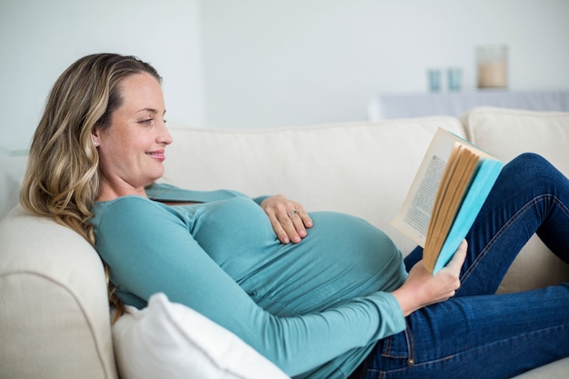 Schwangere Frau, die ein Buch liegt auf der Couch liest