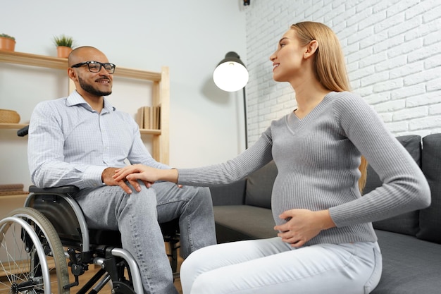 Schwangere Frau, die eine Hand ihres behinderten Mannes hält, der in einem Rollstuhl sitzt