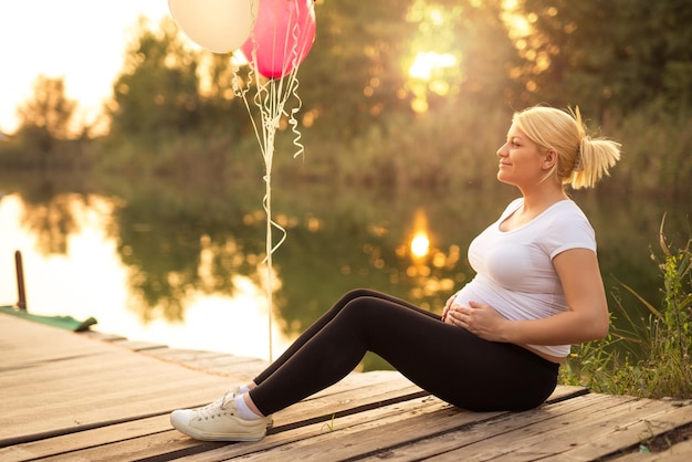 Schwangere Frau, die ihren Bauch hält, während sie neben dem See sitzt