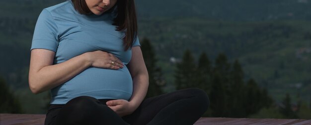 Schwangere Frau, die ihren Bauch umarmt, während sie im Freien sitzt und Yoga mit Kopierraum praktiziert