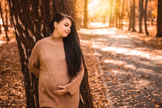 Schwangere Frau, die im herbstlichen Stadtparkwald steht und ihren runden Bauch mit einem kleinen Kind im Inneren streichelt