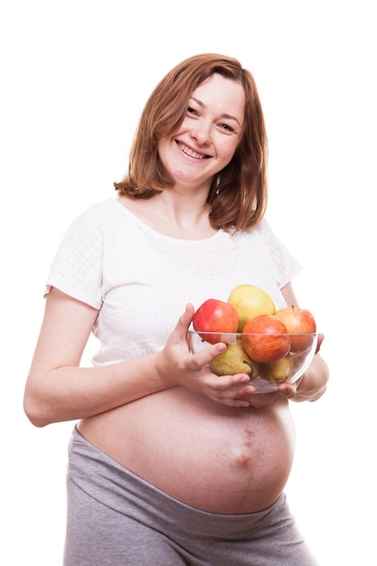 Schwangere Frau, die in die Kamera lächelt, hält eine Glasschale mit Früchten in ihren Händen. Getrennt über weißem Hintergrund
