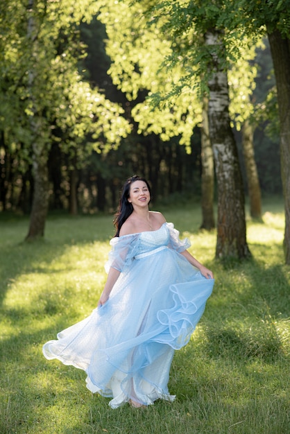 Schwangere Frau, die in einem blauen Kleid auf einem Hintergrund der grünen Bäume aufwirft