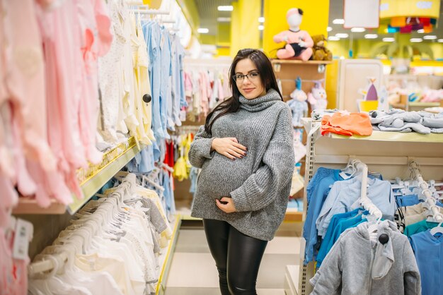 Schwangere Frau, die Kleidung im Laden für Neugeborene kauft. Zukünftige Mutter im Laden von Waren für Säuglinge