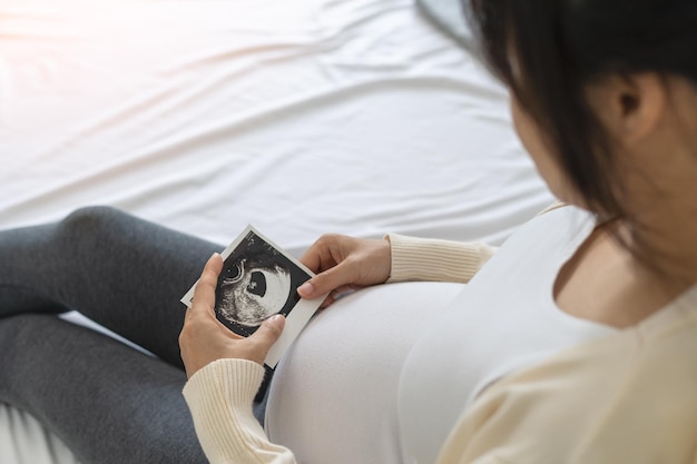 Schwangere Frau, die sich die Ultraschalluntersuchung des Babys ansieht Mama schaut glücklich auf das Ultraschallbild des Babys im Mutterleib auf dem Bett im Haus