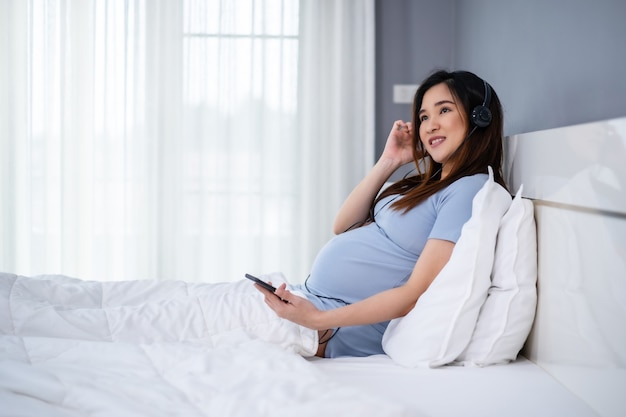 Schwangere Frau, die Smartphone verwendet, um Musik durch Kopfhörer auf einem Bett zu hören