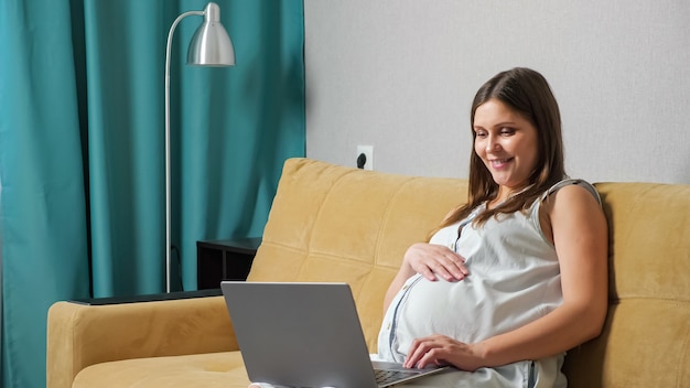 Schwangere Frau, die über einen Videoanruf auf dem Laptop spricht, während sie auf dem Sofa sitzt.