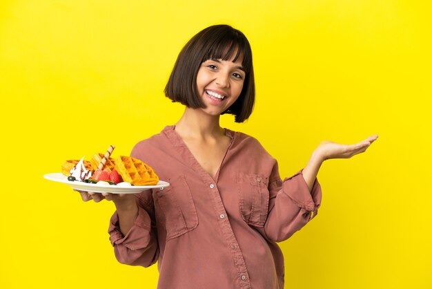 Schwangere Frau, die Waffeln isoliert auf gelbem Hintergrund hält und die Hände zur Seite ausstreckt, um zum Kommen einzuladen