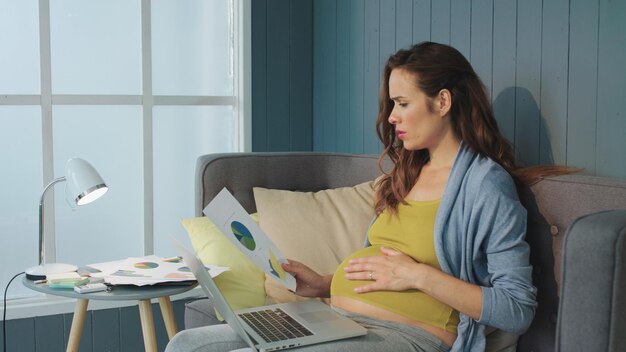 Schwangere Frau, die zu Hause Geschäftsdiagramme analysiert Schwangere Geschäftsfrau, die an einem Remote-Arbeitsplatz arbeitet Abendarbeit Schwangerschaft bei der Geschäftsarbeit Mutter Karriere