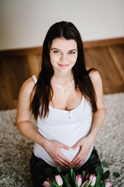Schwangere Frau hält Bauch mit einem Blumenstrauß umarmt Bauch machen Symbol Herz Hände zu Hause Baby-Dusche-Mutterschaft-Konzept Mädchen auf Foto
