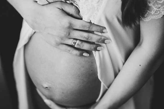 Schwangere Frau hält Hände runden Bauch Mädchen umarmt einen runden Bauch in der Nähe des Fensters zu Hause Mutterschaftskonzept Nahaufnahme neun Monate Babydusche Vorderansicht Schwarz-Weiß-Foto