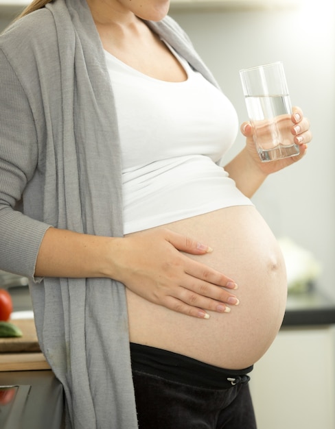 Schwangere Frau im dritten Trimester posiert mit einem Glas Wasser in der Küche