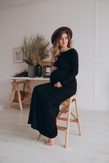 Schwangere Frau im schwarzen Kleid, das auf dem Stuhl im Studio mit weißem Hintergrund sitzt Schwangere Frau, die Bauch berührt