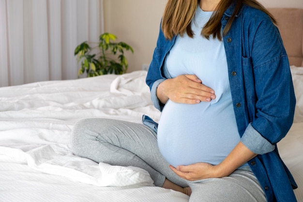 Schwangere Frau in blauem T-Shirt und Jeanshemd, die mit den Händen auf dem Bauch im Bett sitzt Schwangerschaftsmutterschaftsvorbereitungs- und -erwartungskonzept Liebe Babypflege und Elternschaft