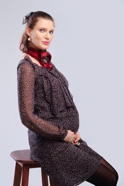 Schwangere Frau in lockerem Kleid sitzt im Profil auf Stuhl auf grauem Studiohintergrund