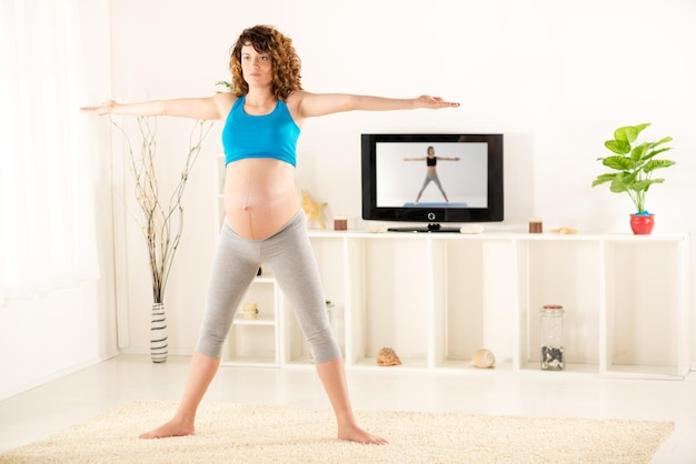 Schwangere Frau in Sportkleidung bei den Übungen im Wohnzimmer, im Hintergrund sieht man den Fernseher, auf dem Übungen gezeigt werden.