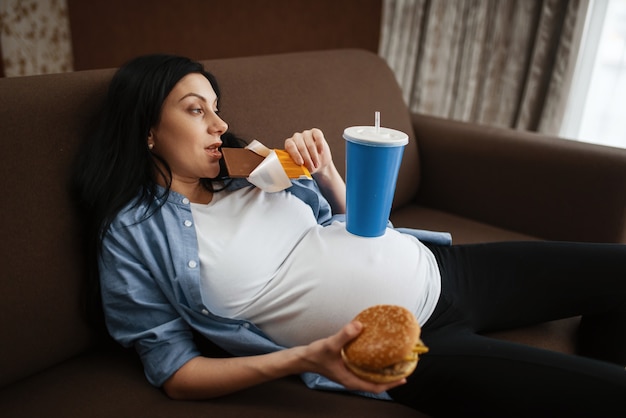 Schwangere Frau mit Bauch trinkt und isst Junk Food zu Hause. Schwangerschaft, Völlerei in der pränatalen Phase. Werdende Mama ruht sich im Wohnzimmer aus