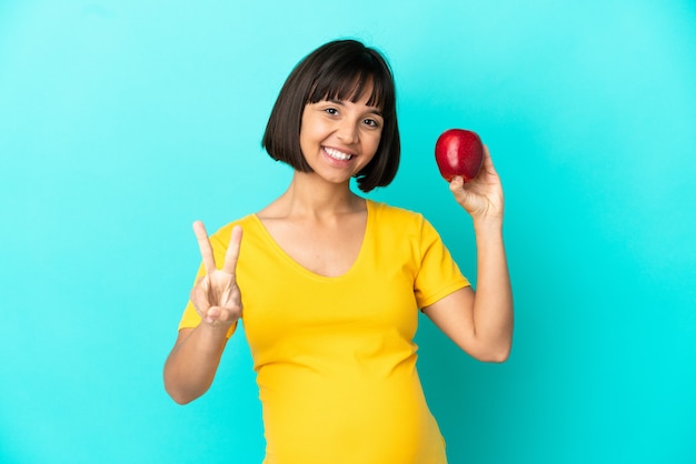 Schwangere Frau mit einem Apfel isoliert auf blauem Hintergrund lächelnd und mit Victory-Zeichen