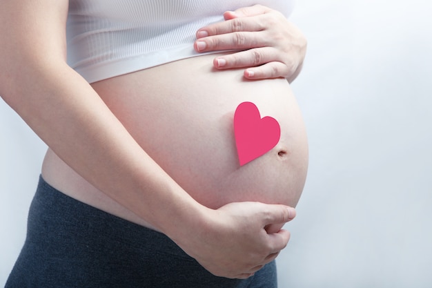 Schwangere Frau mit einzelnem rosa Herz auf ihrem Babybauch. Alles auf weißem Hintergrund.