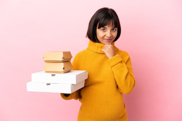 Schwangere Frau mit Pizza und Burger isoliert auf rosa Hintergrund denken