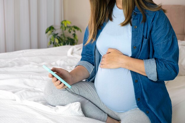 Schwangere Frau mit Smartphone sitzt zu Hause auf dem Bett und nutzt die Schwangerschafts-App, um online zu chatten, in sozialen Medien zu surfen oder Videos anzusehen Mutterschaft und Technologie