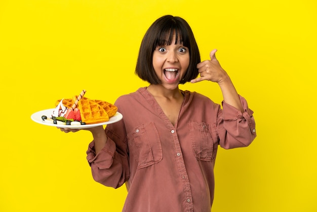 Foto schwangere frau mit waffeln auf gelbem hintergrund isoliert, die telefongeste machen. ruf mich zurück zeichen