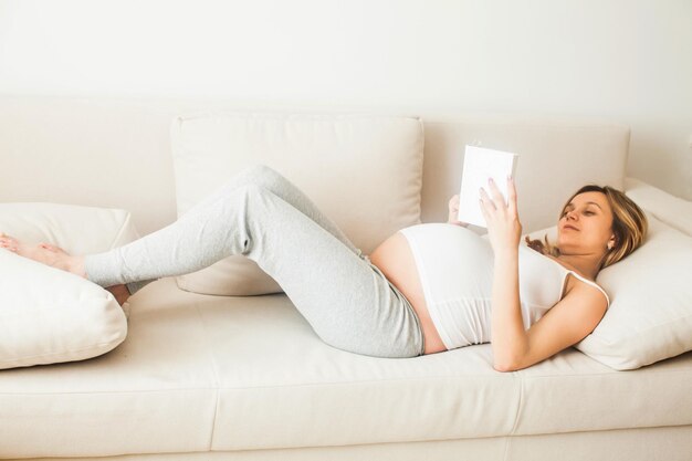 Schwangere Frau ruht sich zu Hause aus und liest Buch auf dem Sofa