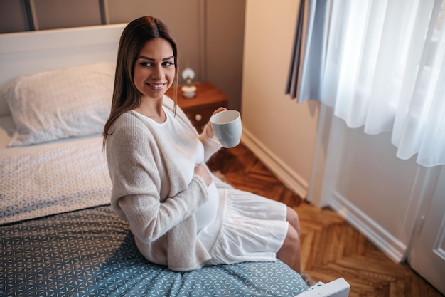 Schwangere Frau sitzt auf dem Bett und trinkt Tee