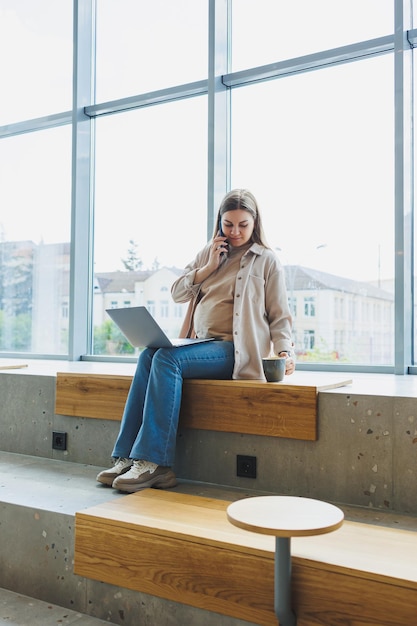 Schwangere Frau sitzt mit Laptop-Smartphone, schaut auf Laptop und macht sich Notizen, während sie im Café sitzt. Eine Frau arbeitet während des Mutterschaftsurlaubs aus der Ferne an einem Laptop