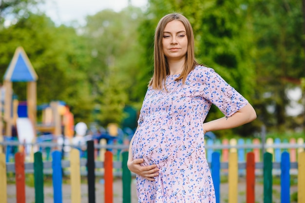 Schwangere Frau steht im Park in der Nähe des Spielplatzes, auf dem Kinder spielen.