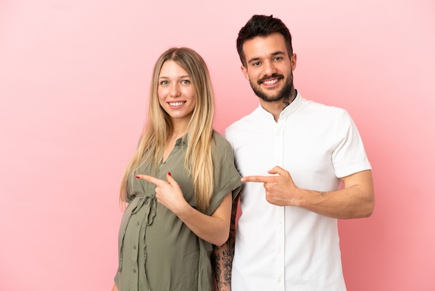 Schwangere Frau und Mann über isoliertem rosa Hintergrund, die mit dem Finger zur Seite zeigen und ein Produkt präsentieren