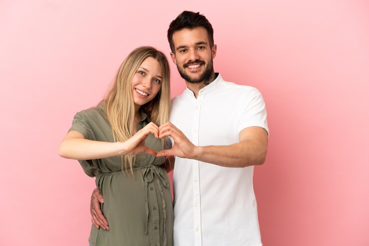 Foto schwangere frau und mann über isoliertem rosa hintergrund machen ein herz mit händen