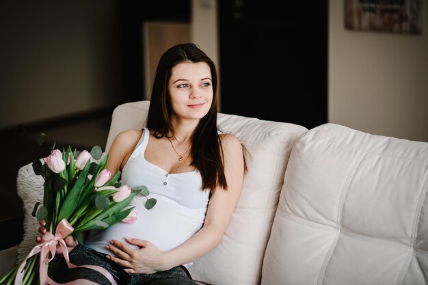 Schwangere, glückliche Frau, die auf dem Bett sitzt und ihren Bauch mit einem Blumenstrauß berührt und zu Hause den Bauch umarmt. Babydusche. Konzept der Mutterschaft. Schwangerschaft, Ruheleute, Erwartungskonzept.