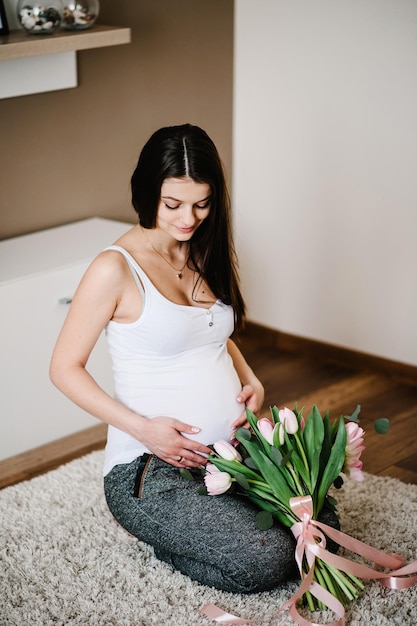 Schwangere glückliche Frau, die auf dem Boden sitzt und ihren Bauch berührt, der einen Blumenstrauß hält, der den Bauch zu Hause umarmt Baby Shower Mutterschaftskonzept Schwangerschaft Rest Erwartung der Menschen