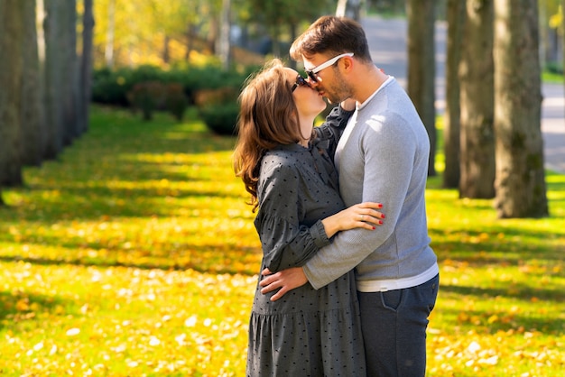 Schwangere junge Frau küsst ihren Ehemann, während sie in einer engen Umarmung draußen in einem bewaldeten Herbstpark stehen