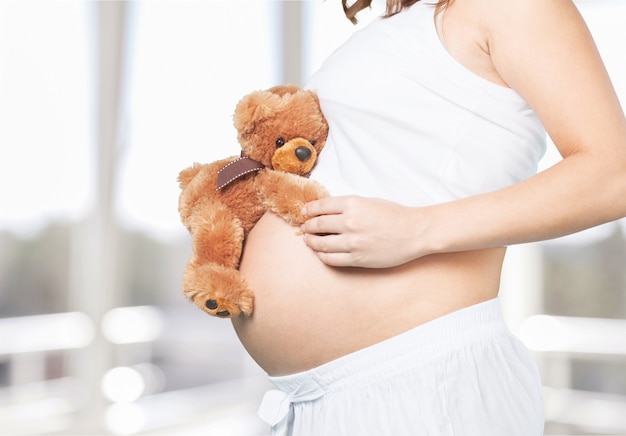 Schwangere junge Frau mit Teddybär auf hellem Raumhintergrund